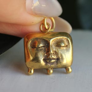 Puede incluir: Un colgante dorado con un diseño de cara cuadrada. La cara tiene los ojos cerrados, una pequeña sonrisa y tres pequeños pies. El colgante está sujeto a un lazo dorado.