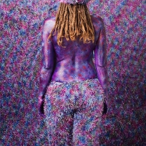 May include: A person with dreadlocks is wearing a fluffy purple hat and trousers. The person is covered in purple, blue and pink glitter paint. The background is a fluffy purple, blue and pink wall.