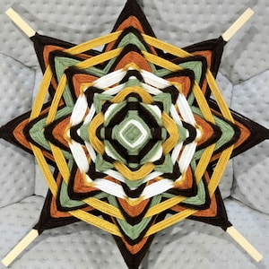 Peut inclure: Une suspension murale en forme d'étoile réalisée avec du fil de laine dans des tons de brun, vert, orange et jaune. Le fil est tissé en un motif circulaire pour créer un design de type mandala.