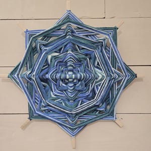 May include: A blue, green, and white yarn mandala with a geometric pattern. The mandala is woven around wooden sticks.