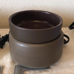 May include: A two-part electric wax warmer in shades of brown. The top bowl is a darker brown, while the base is a lighter, textured brown. The warmer has a power cord and a handle on the side.
