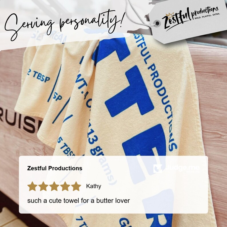 May include: A beige towel with blue text and graphics. The towel features the text "Zestful Productions" and measurements in tablespoons and grams. The towel is folded over a wooden surface.