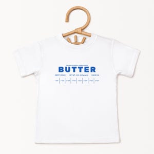 May include: A white t-shirt with the word "BUTTER" in blue block letters. Below the word "BUTTER" are the words "SWEET CREAM", "NET WT. 4 OZ. (113 grams)", and "GRADE AA". The shirt is hanging on a wooden hanger.