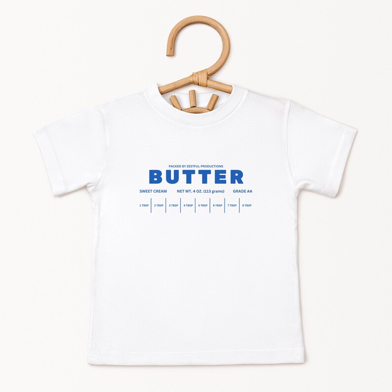 May include: A white t-shirt with the word "BUTTER" in blue block letters. Below the word "BUTTER" are the words "SWEET CREAM", "NET WT. 4 OZ. (113 grams)", and "GRADE AA". The shirt is hanging on a wooden hanger.