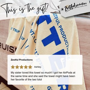 May include: Cream-coloured towel with large blue block letter design and the text "Zestful Productions". The towel also has the text "This is the gift!" in cursive. A customer review is also visible.