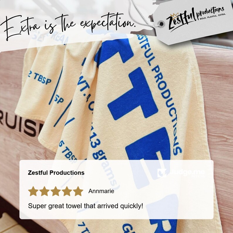 May include: A cream-coloured towel with large blue block letters spelling "ZTF" and the words "Zestful Productions" printed on it. The towel also has text that reads "Extra is the expectation."