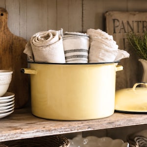 May include: A large, pale yellow enamel stockpot with two side handles, filled with several rolled-up beige and white linen kitchen towels. One towel features thin dark gray stripes. A matching yellow enamel lid sits to the right of the pot.