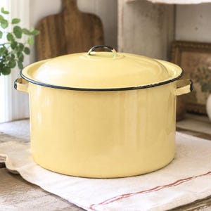 May include: A large, round, pale yellow enamel stockpot with a matching lid. The pot features a black rim along the top edge and two side handles. The surface has a smooth, glossy finish, and the lid includes a small, centered handle knob.
