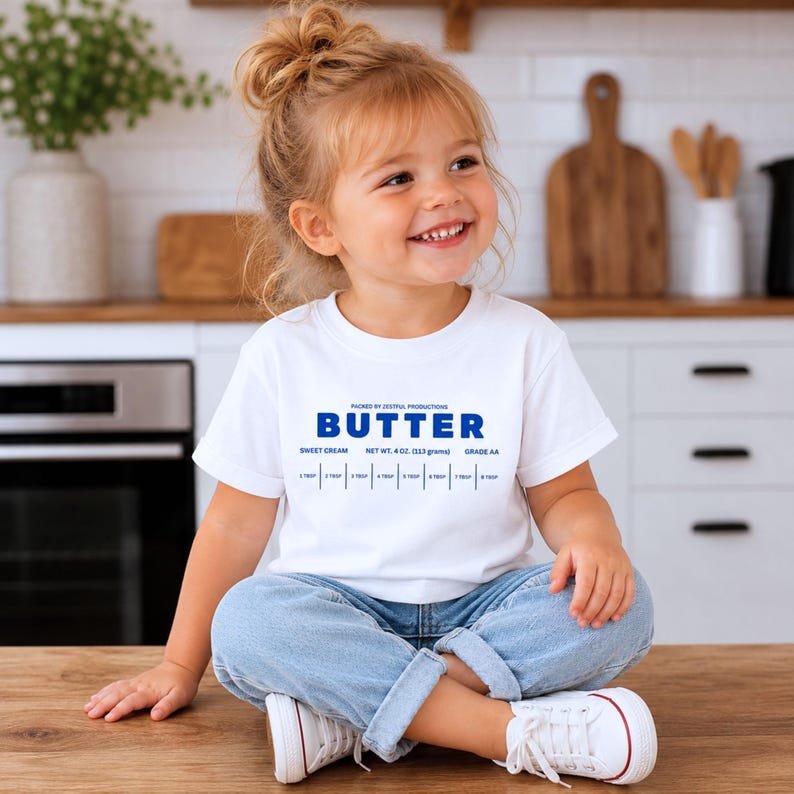 Smiling toddler girl wearing white retro butter label t-shirt with sweet cream foodie graphic. Boutique toddler birthday outfit styled for preschool parties, baking themed celebrations, family photos, and everyday kids fashion with playful vintage charm.
