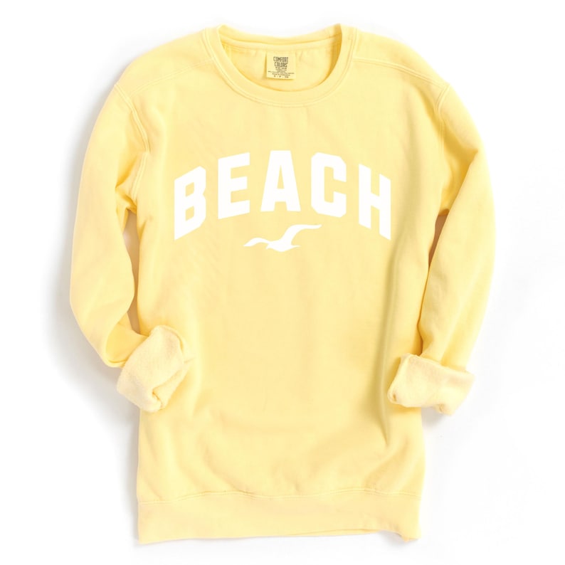 May include: Pale yellow sweatshirt featuring the word "BEACH" in bold white lettering, with a white seagull graphic beneath. The long-sleeved top has a crew neck. The cuffs are rolled up.