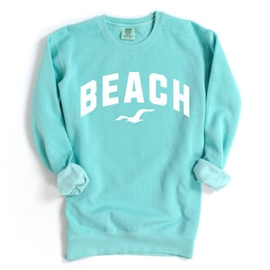 May include: A turquoise sweatshirt featuring the word "BEACH" in bold white lettering. A white seagull graphic is positioned beneath the word. The sweatshirt has a crew neck and long sleeves with rolled cuffs. The background is white.