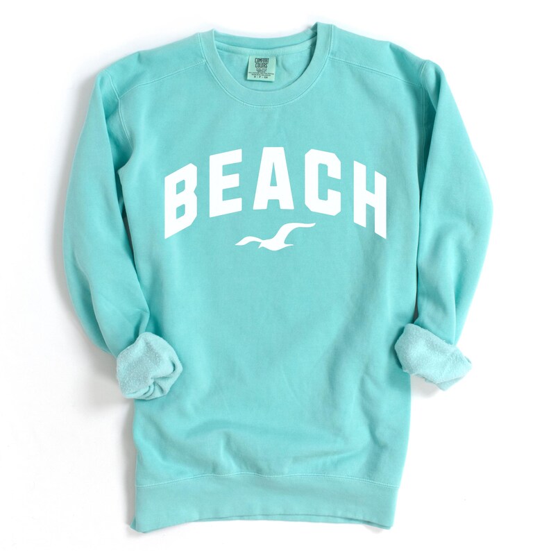 May include: A turquoise sweatshirt featuring the word "BEACH" in bold white lettering. A white seagull graphic is positioned beneath the word. The sweatshirt has a crew neck and long sleeves with rolled cuffs. The background is white.