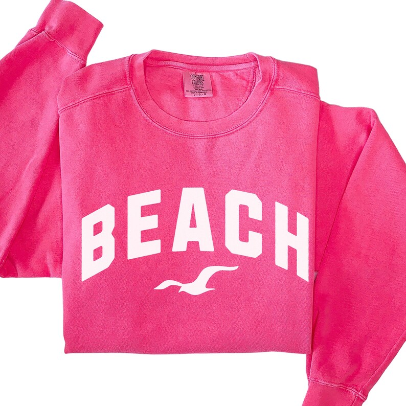 May include: A vibrant pink sweatshirt featuring the word "BEACH" in bold white lettering. Beneath the text is a white seagull design. The sweatshirt has a crew neck and long sleeves.
