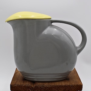 May include: A gray ceramic pitcher with a yellow lid and a curved handle. The pitcher has a rounded body and sits on a wooden surface. The design is modern and sleek, suitable for serving beverages.