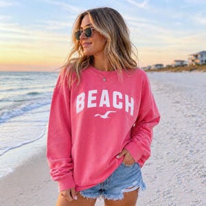 May include: A vibrant coral sweatshirt featuring the word "BEACH" in white, with a seagull graphic. The sweatshirt is paired with denim shorts. The background shows a beach scene with the sea and a sunset.