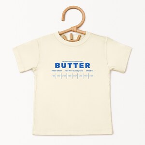 May include: Cream-colored t-shirt with the word "BUTTER" in blue, along with text indicating "SWEET CREAM", "NET WT. 4 OZ. (113 grams)", and "GRADE AA". The shirt is hanging on a wooden hanger.