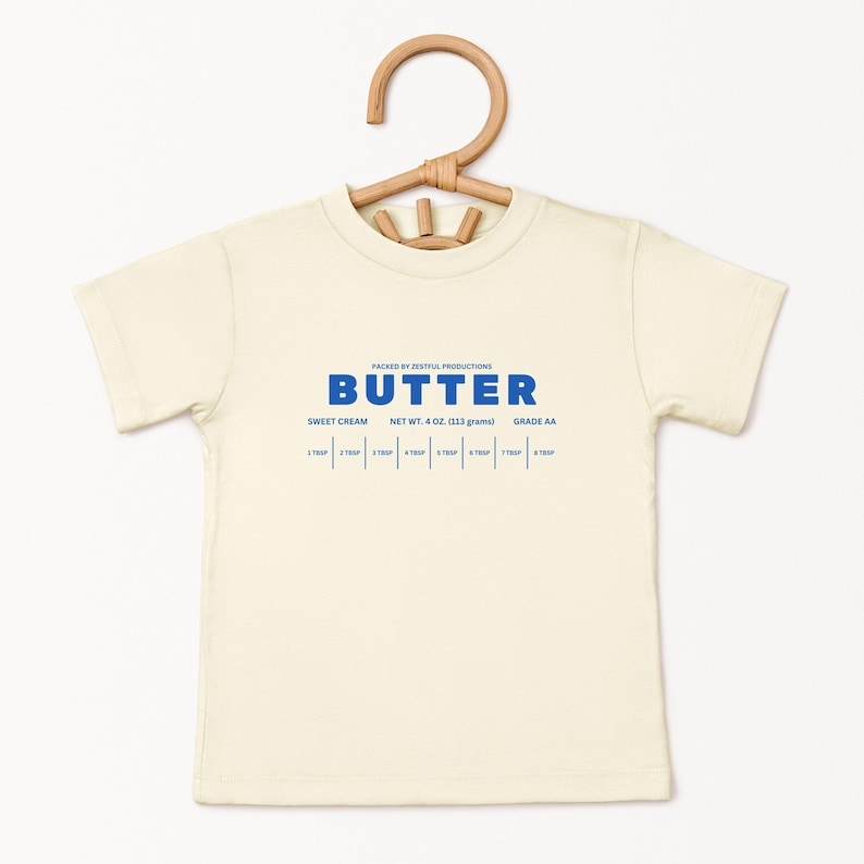 May include: Cream-colored t-shirt with the word "BUTTER" in blue, along with text indicating "SWEET CREAM", "NET WT. 4 OZ. (113 grams)", and "GRADE AA". The shirt is hanging on a wooden hanger.