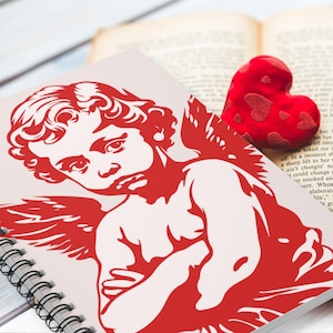 May include: A spiral-bound notebook featuring a red illustration of a cherub with wings. A red heart-shaped decoration with smaller heart patterns rests on an open book. The notebook and book are on a light-colored surface.