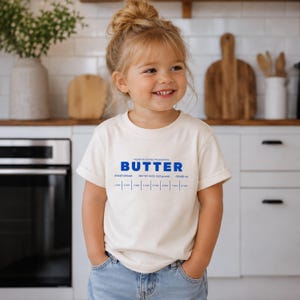Toddler girl wearing cream butter stick label birthday shirt with retro foodie bakery inspired design. Boutique preschool outfit perfect for birthday parties, baking themed celebrations, stylish family photos, and playful everyday kidswear.