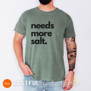 Man wearing moss green ‘Needs More Salt’ shirt with bold black text – sarcastic gift idea for chefs and cooks.