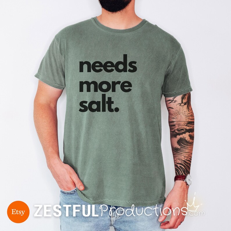 Man wearing moss green ‘Needs More Salt’ shirt with bold black text – sarcastic gift idea for chefs and cooks.