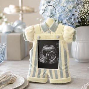 May include: A baby-themed photo frame shaped like a onesie, in soft yellow with blue stripes and accents. The frame holds a black and white ultrasound image. The frame has a hanger at the top, suggesting it can be displayed.