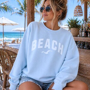 May include: A pale blue sweatshirt featuring the word "BEACH" in white, with a seagull graphic. The sweatshirt is paired with denim shorts. The background shows a beach with palm trees and parasols, suggesting a coastal theme.
