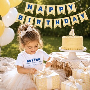 Toddler birthday girl wearing butter themed retro kids t-shirt at luxury outdoor picnic party with matching pennant banner and birthday cake. Boutique foodie toddler outfit styled for birthday celebrations, cake smash photos, and playful baking themes.