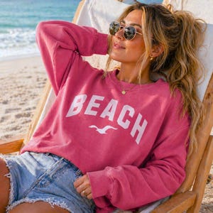 May include: A pink sweatshirt with the word "BEACH" in white block letters and a white seagull graphic. The sweatshirt is worn with denim shorts. The person is wearing sunglasses and gold jewellery.