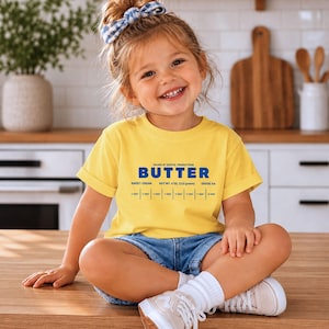 Smiling toddler girl wearing retro butter stick label toddler shirt in cozy boutique kitchen setting. Cute foodie kids tee with sweet cream baking design styled as trendy birthday girl outfit for preschool parties, family photos, and playful everyday wear.