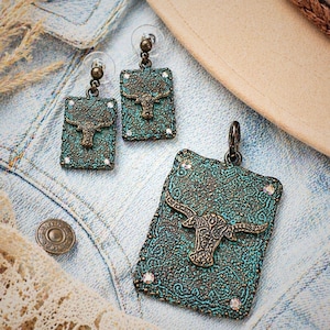 May include: A turquoise and bronze jewelry set featuring a pendant and matching earrings. Each piece has a rectangular shape with a textured pattern and a bull head design. The set includes small, clear crystals.