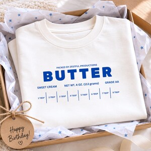 Folded white butter stick label toddler shirt featuring retro sweet cream baking graphic and foodie inspired design. Boutique preschool party apparel styled with soft neutral packaging for trendy birthday outfits, baking lovers, and kids fashion photography.