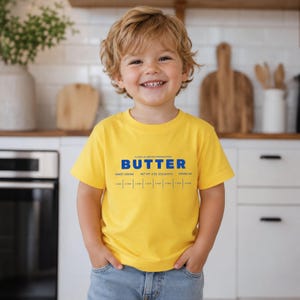 Little boy wearing yellow butter toddler shirt with retro baking packaging graphic in modern kitchen setting. Soft foodie kids tee designed for playful preschool outfits, baking families, birthday parties, and trendy boutique children’s apparel.