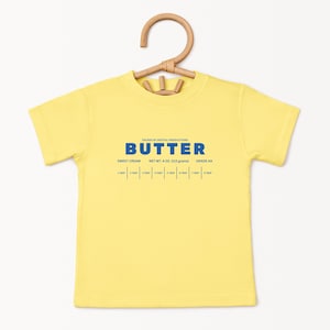 May include: A yellow short-sleeved t-shirt with the word "BUTTER" in blue block letters. Below the word "BUTTER" are the words "SWEET CREAM", "NET WT. 4 OZ. (113 grams)", and "GRADE AA". The shirt is hanging on a wooden hanger.