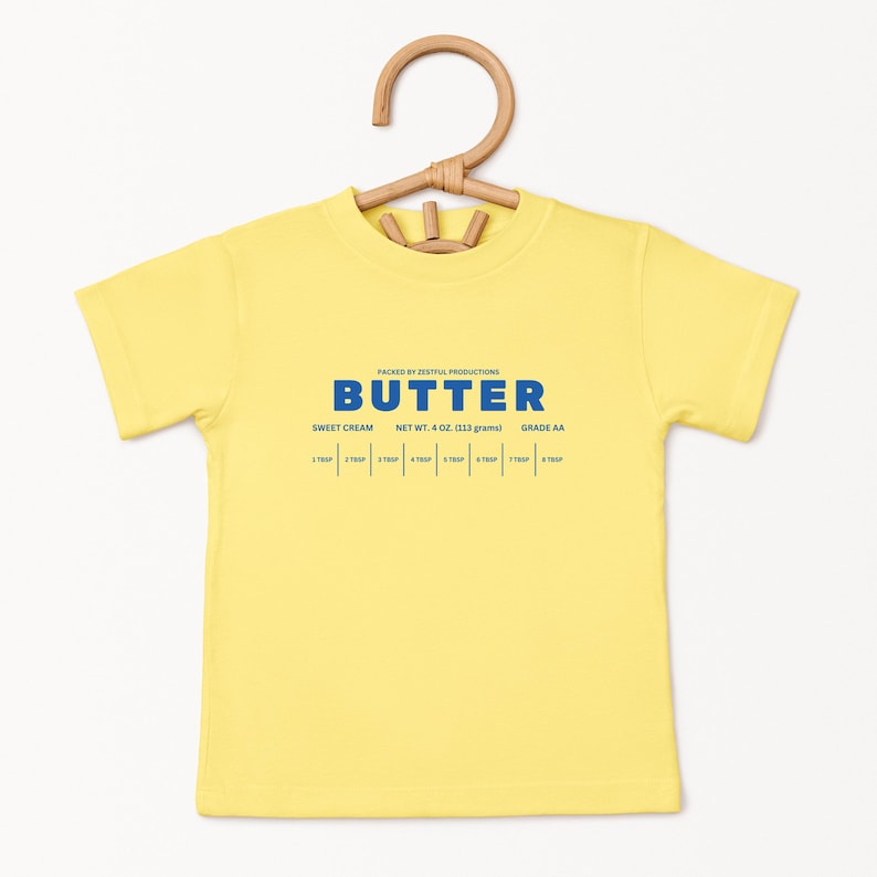 May include: A yellow short-sleeved t-shirt with the word "BUTTER" in blue block letters. Below the word "BUTTER" are the words "SWEET CREAM", "NET WT. 4 OZ. (113 grams)", and "GRADE AA". The shirt is hanging on a wooden hanger.
