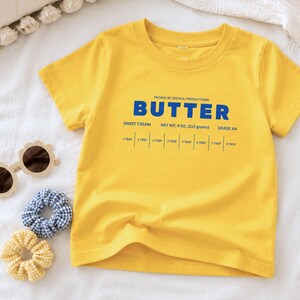 Yellow butter toddler shirt flat lay with retro sweet cream baking design and foodie inspired packaging graphic. Boutique preschool apparel styled for birthday parties, baking lovers, kitchen helper outfits, and trendy children’s boutique fashion.