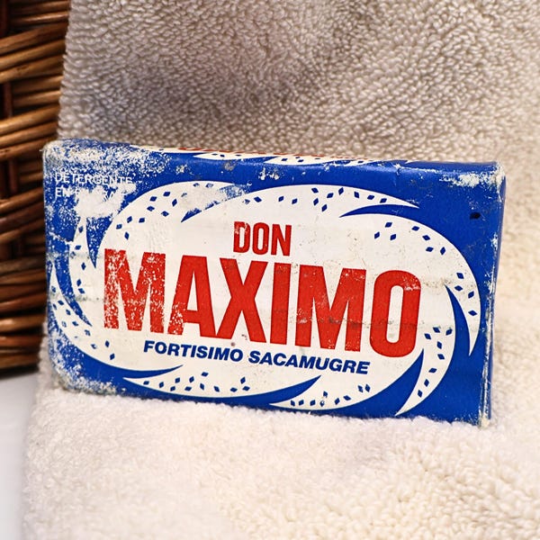 May include: A rectangular bar of Don Maximo detergent soap in a blue and white paper wrapper. The front features the red text DON MAXIMO, with the blue text FORTISIMO SACAMUGRE below it, surrounded by a white oval graphic with blue accents.