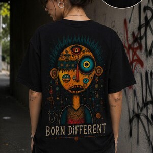 May include: Black t-shirt featuring a colorful, stylized illustration of a face with multiple eyes and the text "BORN DIFFERENT". The shirt also has a small eye design on the front. The person is wearing black jeans.