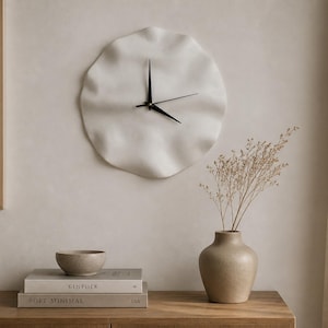 May include: A white, wavy-edged wall clock with black hands hangs above a wooden cabinet. A small vase holds dried flowers, and a bowl and books are on the cabinet. The room has a neutral colour palette.
