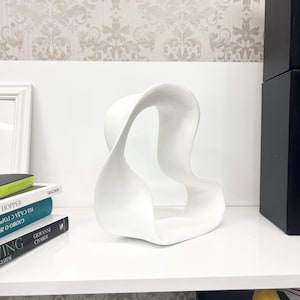 Puede incluir: Una escultura abstracta blanca con un diseño fluido en forma de cinta. La escultura está hecha de un material liso y sólido y se encuentra sobre una superficie blanca. El fondo presenta un papel pintado estampado y libros.