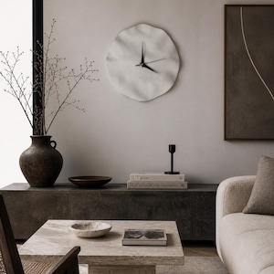 May include: A contemporary living room with a white, organic-shaped wall clock with black hands. A dark brown vase holds branches. A stone coffee table displays a bowl and a book. A sofa and a cabinet complete the decor.