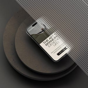 May include: A smartphone displaying a screen with the text ROWAN, THE ART OF BRANDING (DESIGNER LIFE), and START COURSE, partially obscured by a ribbed glass panel, resting on two stacked dark gray circular platforms against a dark background.