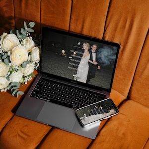 May include: A laptop and smartphone displaying a wedding website are placed on an orange velvet couch. A bouquet of white roses and greenery rests beside the laptop. The laptop screen shows a couple in formal attire. The smartphone mirrors the laptop's display.