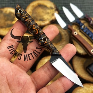 May include: A small, decorative knife with a patterned handle in shades of brown and orange. The blade is silver, and the handle has three circular inlays. The knife is held in a hand, with other knives visible in the background.