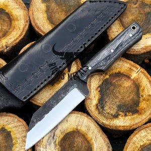 May include: A black leather sheath with a decorative pattern and a black handled knife with a silver blade. The knife is resting on a pile of wooden rounds.