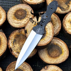 Peut inclure: Un poignard noir et argent avec une poignée noire et une lame argentée. Le poignard repose sur une pile de rondins de bois. Le texte "Rick's Metals" est visible sur la poignée.