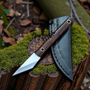 May include: A handcrafted knife with a dark wooden handle and a silver blade, resting on a bed of green moss. The knife is accompanied by a black leather sheath. The handle has two metal rivets. The knife is a hunting or utility tool.