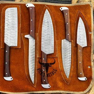 Peut inclure: Ensemble de cinq couteaux de cuisine avec des manches en bois, pr&eacute;sent&eacute;s dans un rouleau en cuir brun avec le texte "Rick's Metals" sur le devant.