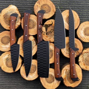 Peut inclure: Cinq couteaux de cuisine avec des lames noires et des manches en bois sont dispos&eacute;s sur un fond de rondins de bois empil&eacute;s. Les lames ont une finition martel&eacute;e et les manches sont de couleur brun fonc&eacute;. Les couteaux sont dispos&eacute;s en rang&eacute;e, les lames pointant vers le haut de l'image. Le texte "Rick's Metals" est visible sur l'un des couteaux.