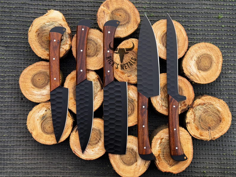 Peut inclure: Cinq couteaux de cuisine avec des lames noires et des manches en bois sont dispos&eacute;s sur un fond de rondins de bois empil&eacute;s. Les lames ont une finition martel&eacute;e et les manches sont de couleur brun fonc&eacute;. Les couteaux sont dispos&eacute;s en rang&eacute;e, les lames pointant vers le haut de l'image. Le texte "Rick's Metals" est visible sur l'un des couteaux.